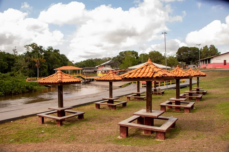 Mesas com cobertura de telhas na orla de Mazagão Velho, com o rio, passarelas e mata ao fundo sob céu azul com nuvens.