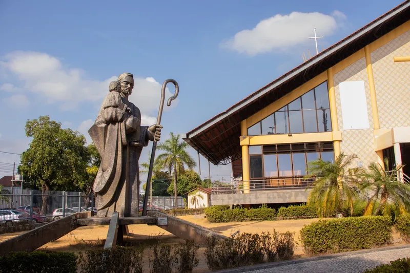 Gran estatua religiosa con cayado frente a la fachada acristalada de la Catedral de São José de Macapá, bajo cielo azul.
