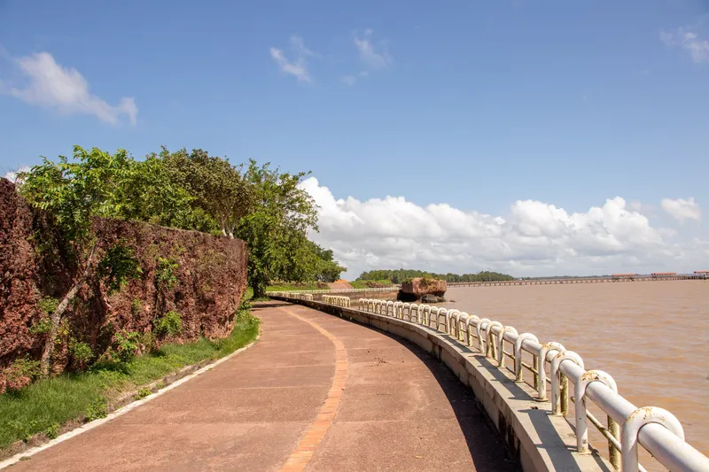 Promenade courbe du Parque do Forte le long du mur de pierre rougeâtre, avec garde-corps blanc au bord du fleuve et ciel bleu nuageux.