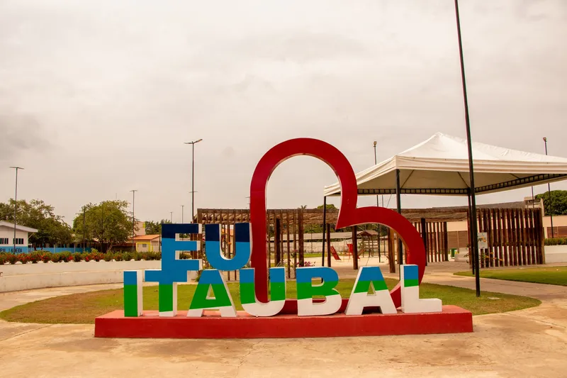 Letreiro colorido "Eu ♥ Itaubal" na praça, com tenda branca e pergolado de madeira ao fundo
