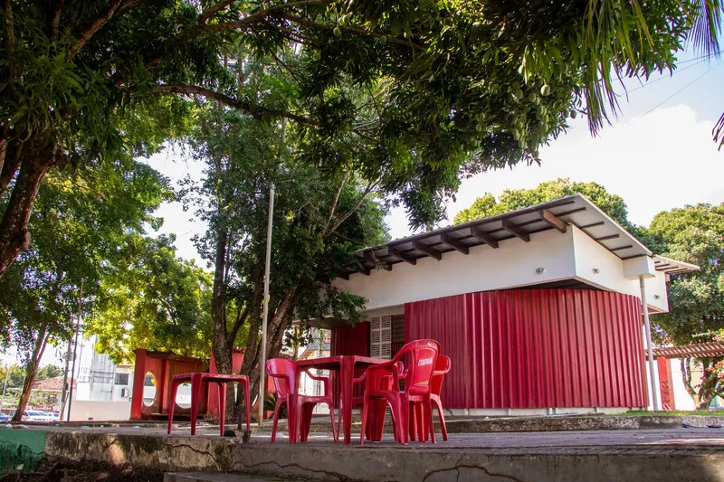 Mesa e cadeiras vermelhas diante de um quiosque branco com paredes vermelhas sob árvores na Praça Isaac Zagury, vistos de ângulo baixo.