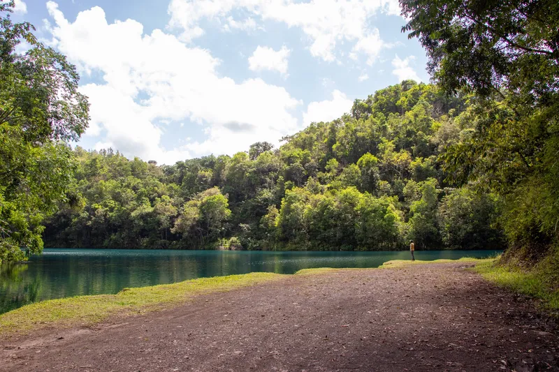 Dirt shore of the Lagoa Azul in Serra do Navio, with blue-green water and a forested hill under a cloudy sky.