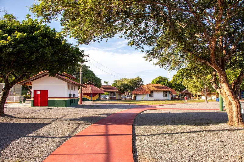 Caminho vermelho entre áreas de pedrisco levando a casinhas brancas com telhado de cerâmica no Complexo do Araxá, sob árvores.