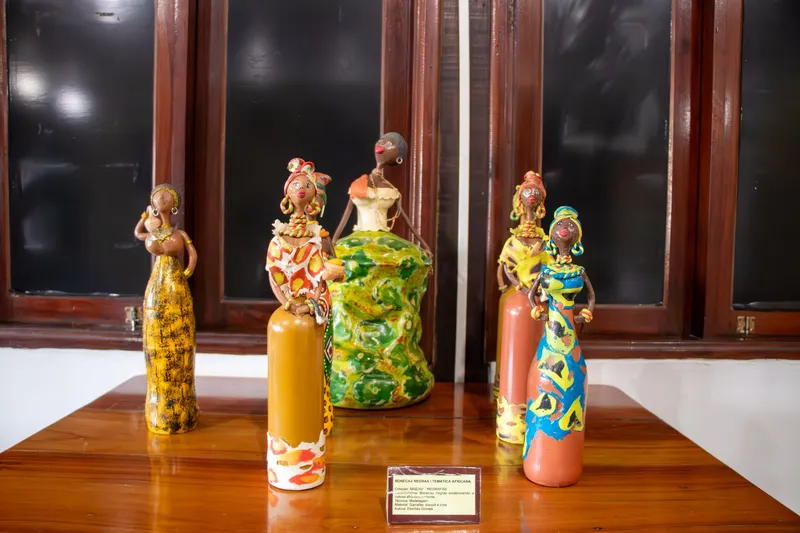 Bonecas de cerâmica pintadas com temática africana, com turbantes e vestidos coloridos, expostas sobre mesa de madeira no Museu de Arqueologia e Etnologia.