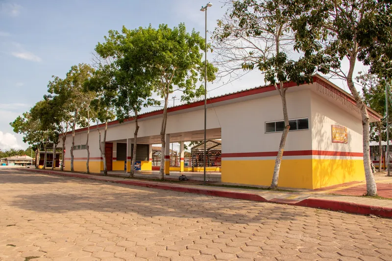 Long kiosk building in the Mazagão Novo square, painted white, yellow and red, with trees along the sidewalk.