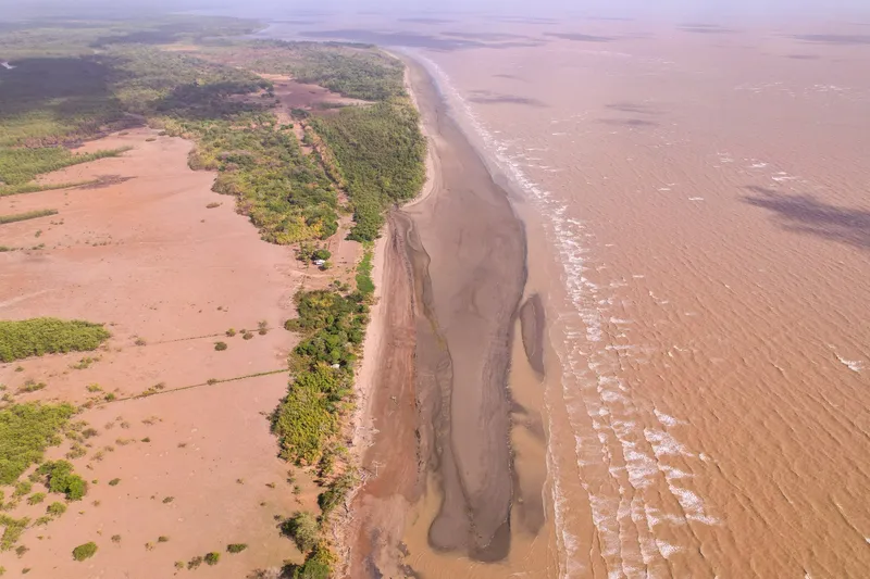 Vista aérea vertical do litoral da Praia do Goiabal: mar barrento com ondas, banco de areia e lama, e faixa de vegetação ao lado do campo