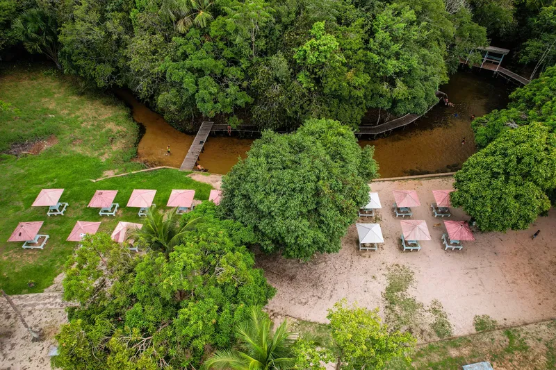 Aerial view of Balneário São Joaquim do Pacuí: pink-roofed kiosks on sand and grass, the creek and boardwalks in the forest.
