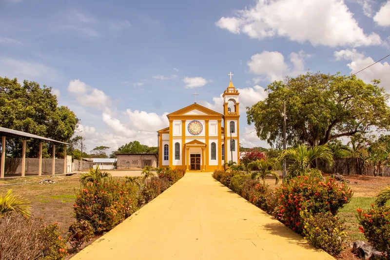 Caminho amarelo ladeado de canteiros floridos levando à fachada branca e amarela da Igreja Nossa Senhora de Nazaré, sob céu azul com nuvens