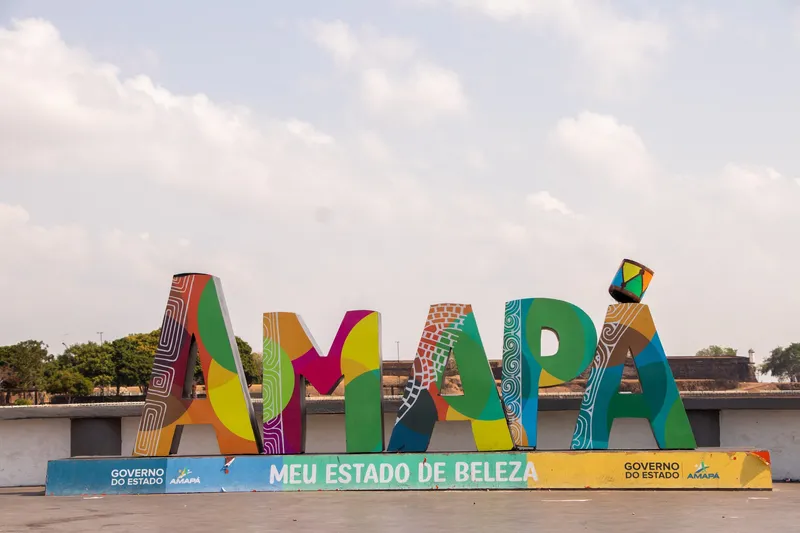 Letrero colorido "Amapá, meu estado de beleza" con grafismos en la orla, bajo un cielo claro, con la muralla de la fortaleza y árboles al fondo.