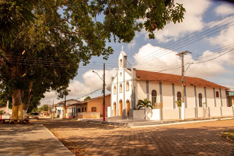 Igreja Nossa Senhora da Assunção on the corner of a paved street in Mazagão Velho, with a large tree and houses alongside.