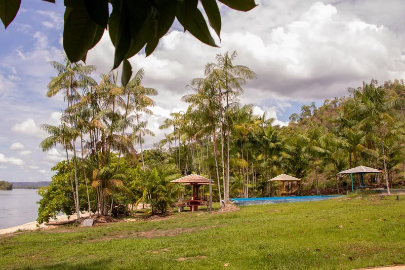 Lawn with covered kiosks, a swimming pool and açaí palms at Balneário Belas Águas, with the river on the left and a cloudy sky.