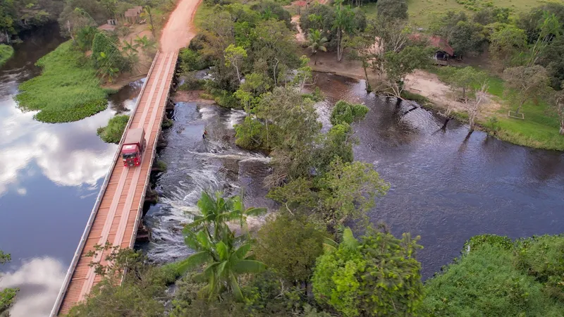 Vista aérea del Balneário Água Fria: puente de madera con un camión sobre el río, rápidos al lado y selva en las orillas