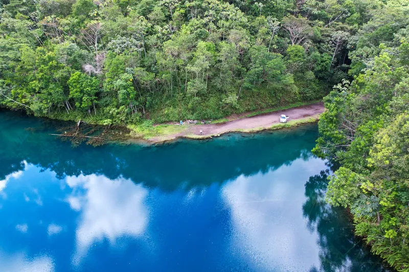 Aerial view of the Lagoa Azul in Serra do Navio, deep blue water reflecting clouds, surrounded by forest with a shoreline road.