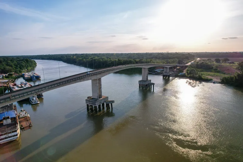 Aerial view of the Ponte da Integração over the Matapi River at dusk, with sun reflecting on the water and moored barges.
