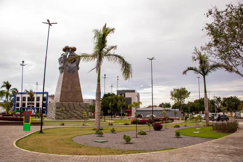 Monument of Praça Povos do Meio do Mundo seen from street level, with palm trees, lawn and a gravel bed under a cloudy sky.
