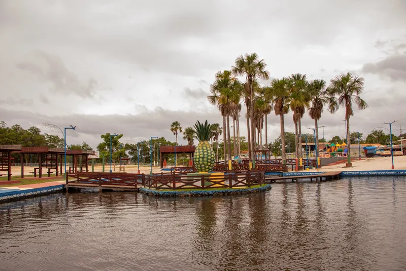 Escultura de piña gigante en la isla central del lago del balneario de Porto Grande, con cerca de madera, palmeras y cielo nublado