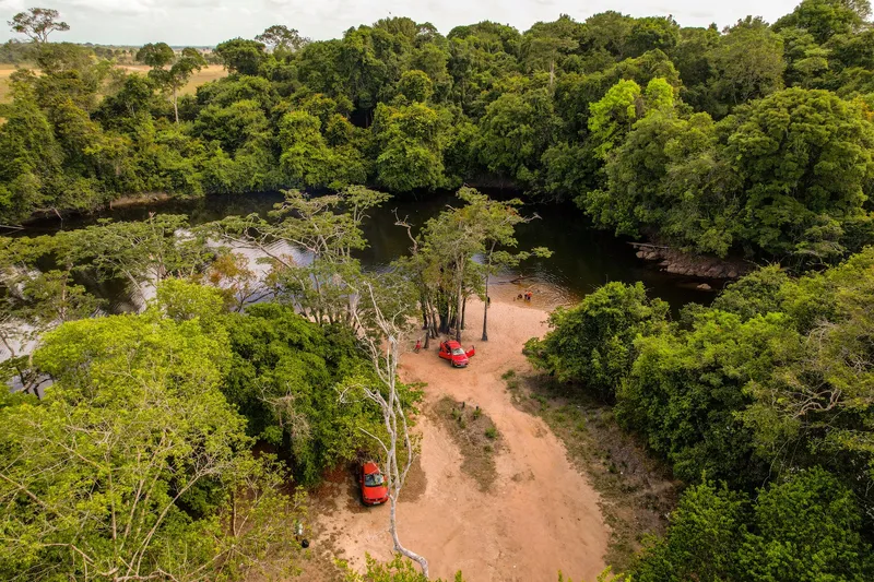 Aerial view of Balneário da Prainha: a sandy clearing with parked cars beside the dark river, surrounded by dense forest