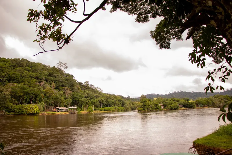 Wide, calm Amapari River seen from the bank under tree branches, with houses and forested hills under a cloudy sky.