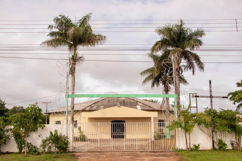 Fachada da Igreja Nossa Senhora do Perpétuo Socorro, prédio amarelo de telhado baixo atrás de portão gradeado, entre duas palmeiras altas.