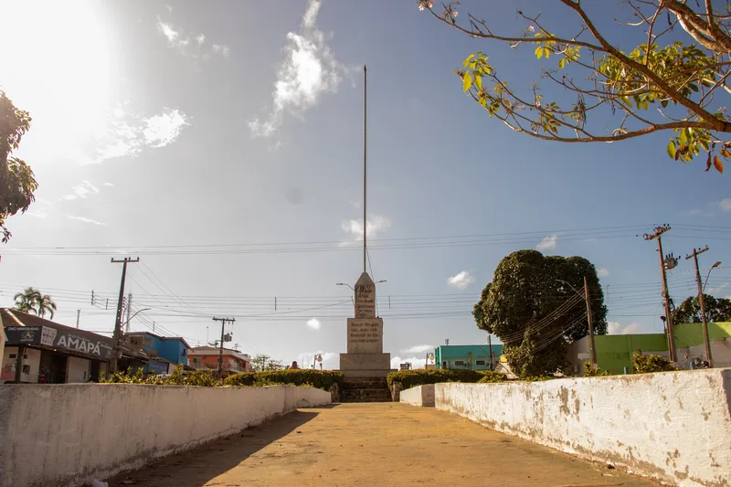Camino entre muretes blancos que lleva al obelisco del Monumento a la Patria, con mástil alto, sol intenso y casas de Oiapoque alrededor