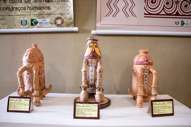 Três réplicas de urnas funerárias antropomorfas em cerâmica, com placas explicativas, expostas sobre mesa branca no Museu de Arqueologia e Etnologia.