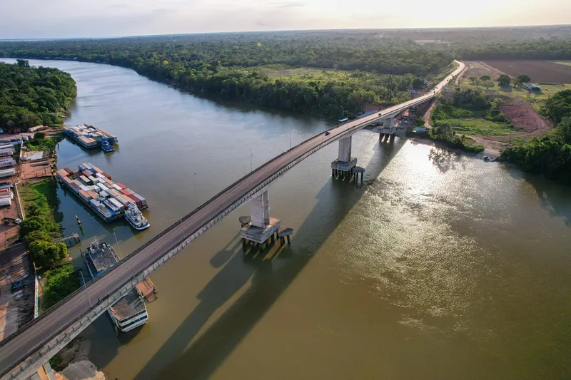 The Ponte da Integração crossing the Matapi River seen from above in late afternoon, with container barges on the bank and forest.