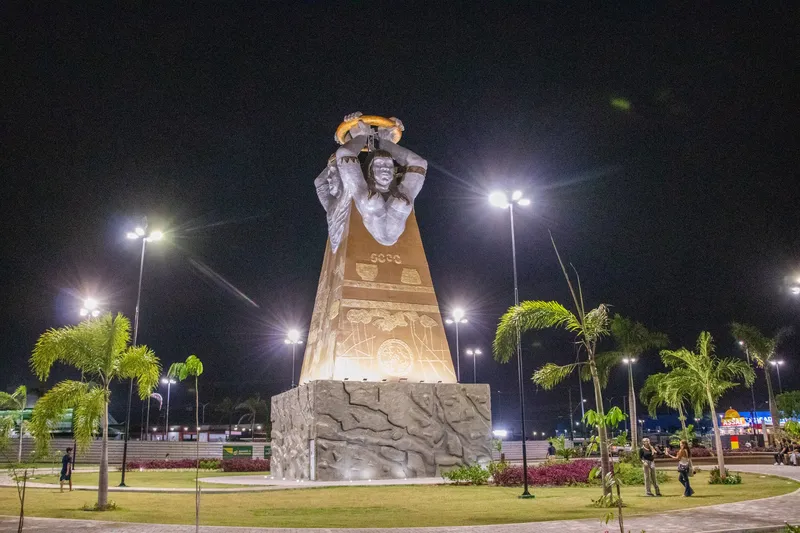 Monument of Praça Povos do Meio do Mundo lit up at night, with glowing street lamps, palm trees and lawn around it.