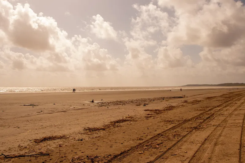 Faixa larga de areia da Praia do Goiabal com marcas de pneu, mar ao fundo, pessoas pequenas ao longe e céu com grandes nuvens