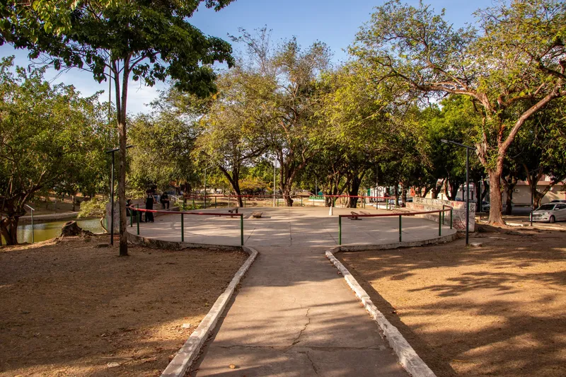 Caminho de concreto levando a uma área circular cercada por árvores na Praça Floriano Peixoto, com o lago à esquerda e luz de fim de tarde.
