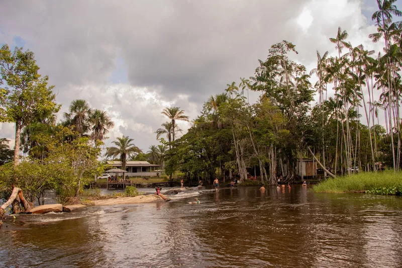 Bañistas pequeños a lo lejos en el río de aguas oscuras del Balneário Água Fria, con palmeras, casas y cielo cargado