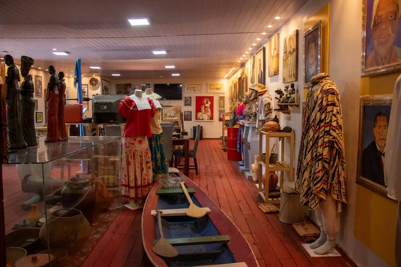 Salão do Museu Afro-Amazônico Josefa Pereira Laú com piso de madeira vermelha, uma canoa com remos, manequins vestidos, esculturas e quadros.