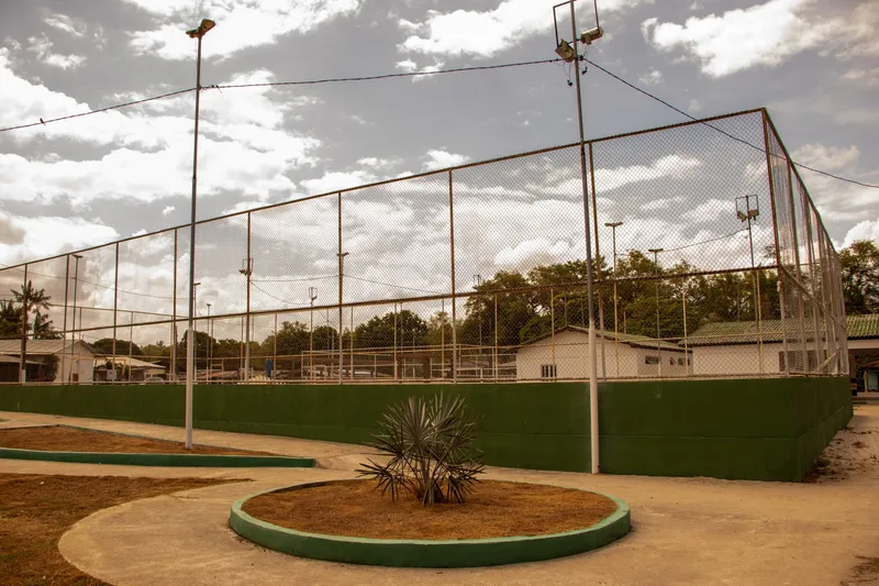 Cancha deportiva rodeada por alambrado alto y muro bajo verde en la Praça Cabralzinho, con jardinera redonda y postes de iluminación