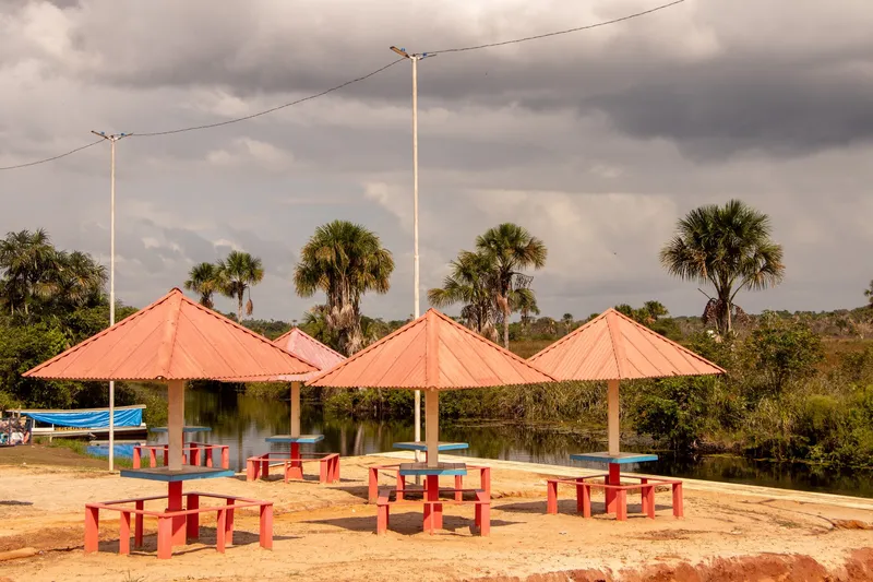 Quiosques de telhado laranja com mesas e bancos no Balneário Gurupora, à beira do rio com buritizeiros e céu carregado ao fundo