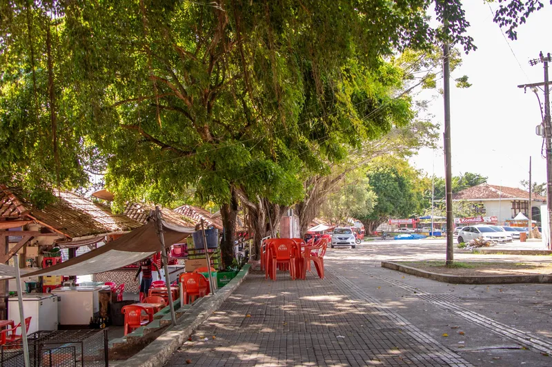 Calçada sombreada da Praça Isaac Zagury com quiosques de telhado de palha e cadeiras vermelhas sob grandes árvores.