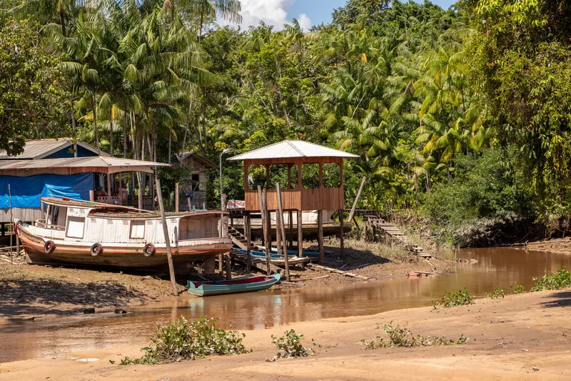 Rive de la rivière Paxicu avec un bateau en bois posé sur le sable, une pirogue, une maison sur pilotis et des palmiers açaí au fond.
