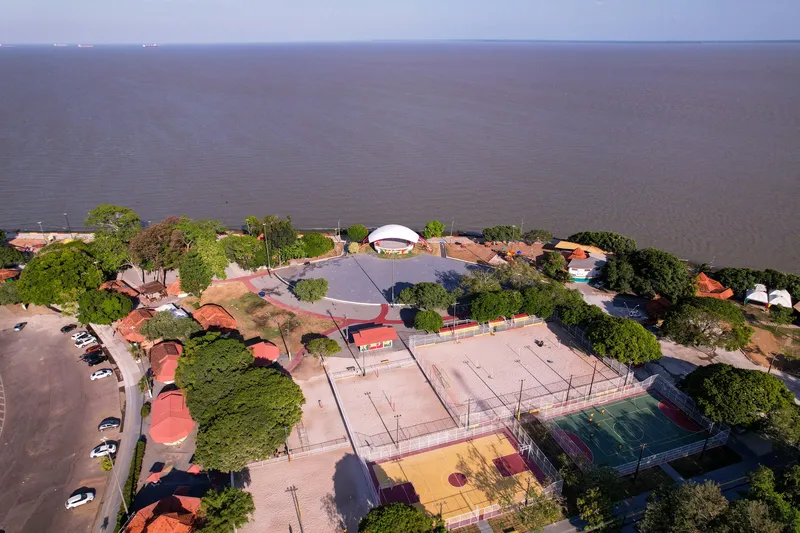 Vista aérea do Complexo do Araxá com quadras esportivas, praça com concha acústica e quiosques à beira do rio Amazonas, no fim da tarde.