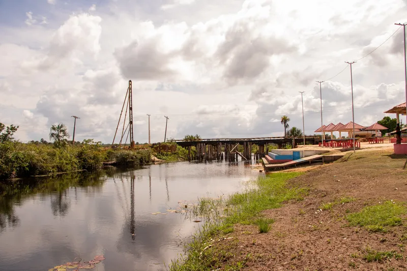 Rio do Balneário Gurupora com ponte de madeira ao fundo, deque colorido e quiosques na margem gramada, sob céu nublado