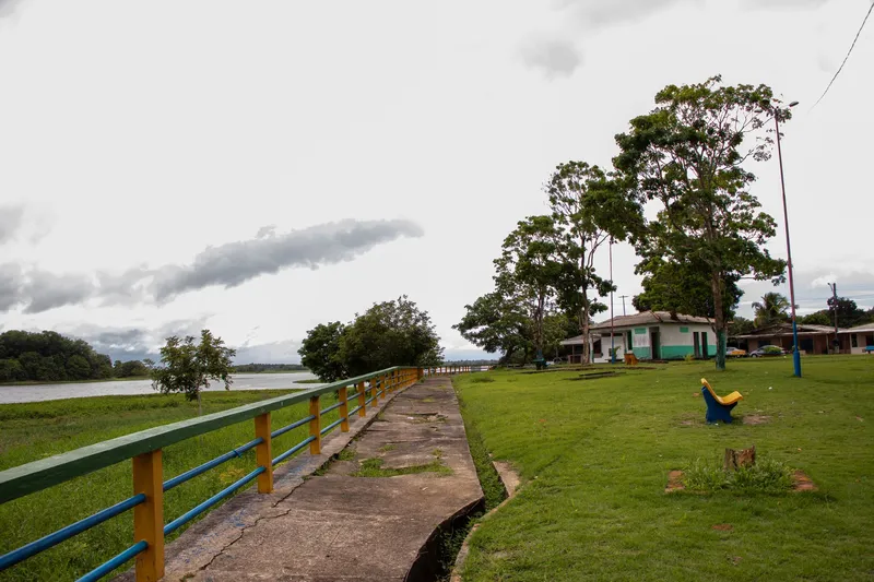 Calçada da Orla de Pracuúba com guarda-corpo verde, amarelo e azul, gramado com árvores e o rio à esquerda.