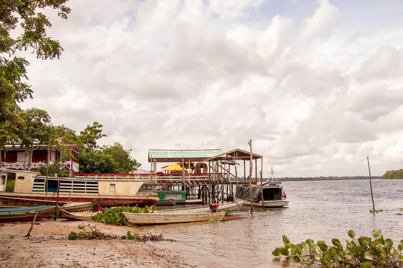 Rive sablonneuse du front de rivière de Cutias do Araguari avec pirogues, bateaux en bois et ponton couvert, large fleuve et ciel couvert