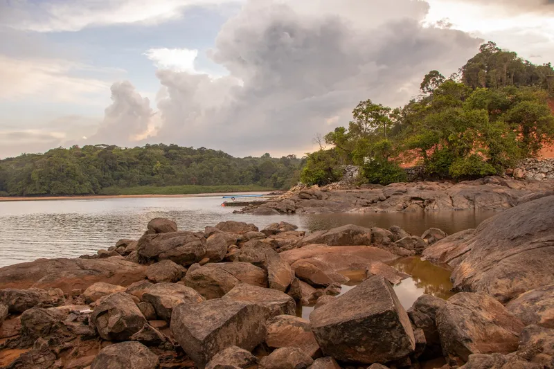 Rochas avermelhadas em primeiro plano na margem do rio Oiapoque, com um barco atracado, mata ao fundo e nuvens altas ao fim da tarde