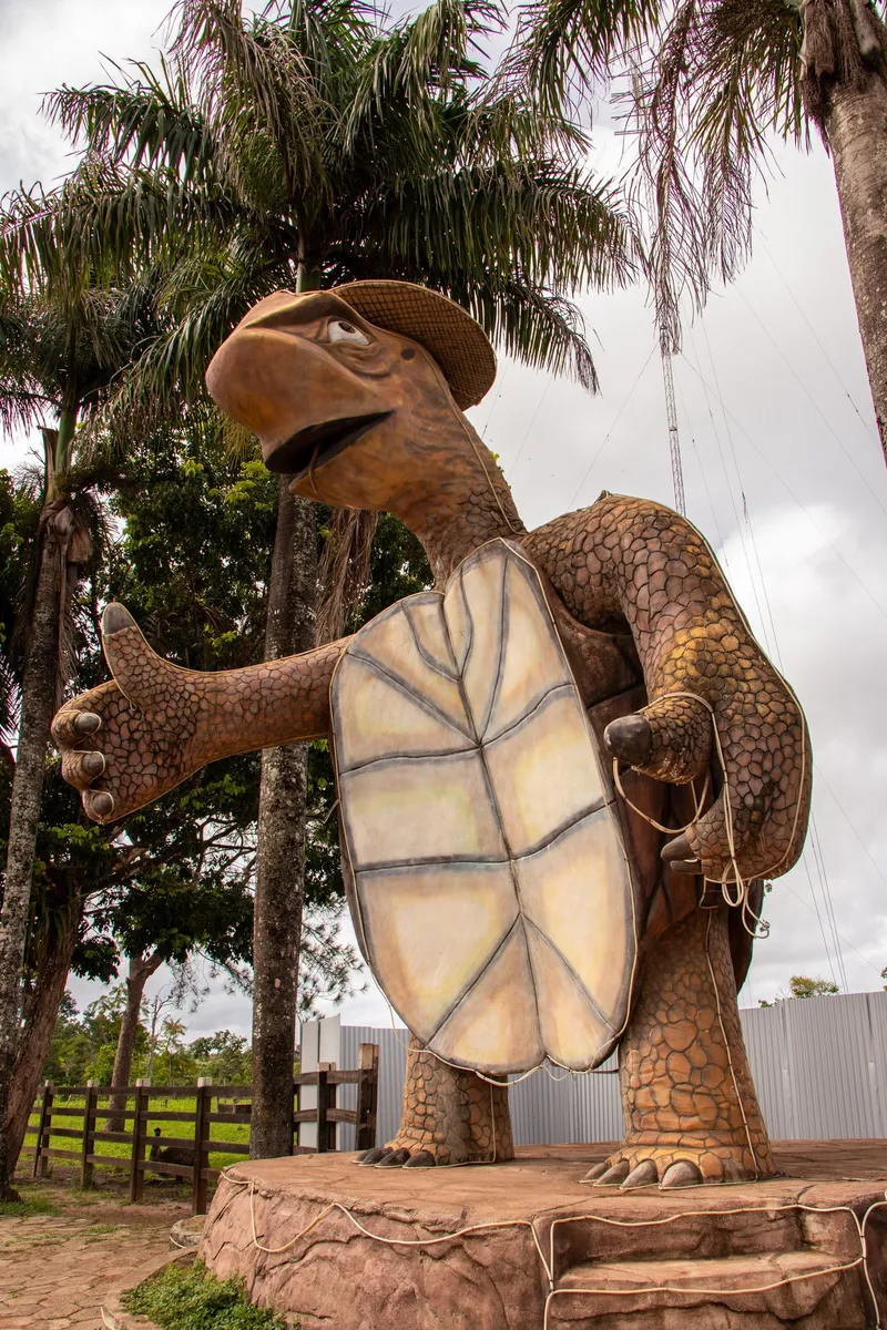 Monumento da Tartaruga visto desde abajo, en formato vertical: la tortuga con sombrero y el pulgar levantado, rodeada de palmeras.