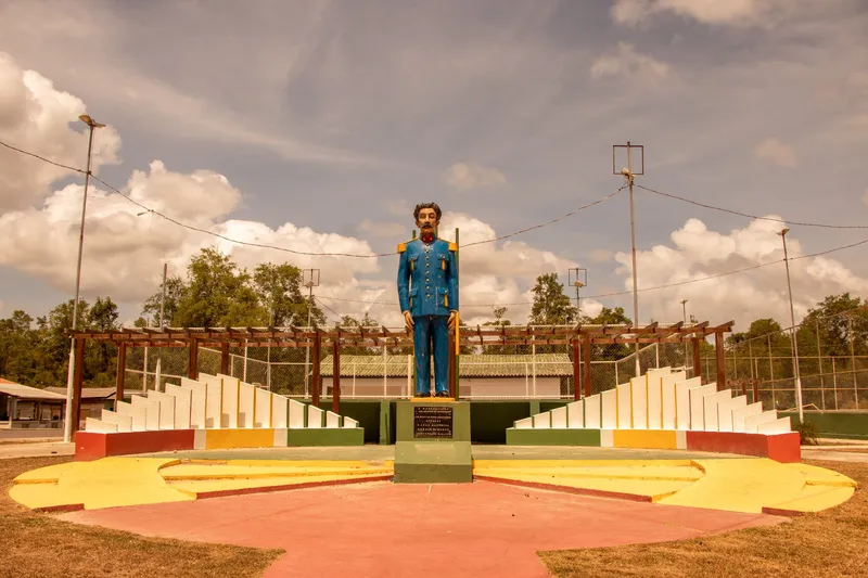 Estatua colorida de un hombre con uniforme azul sobre pedestal en la Praça Cabralzinho, flanqueada por estructuras escalonadas y pérgolas, con piso amarillo, rojo y verde