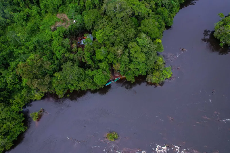 Vista aérea cenital de un río oscuro con rápidos, orilla de selva con pequeña construcción y botes atracados en Tumucumaque.