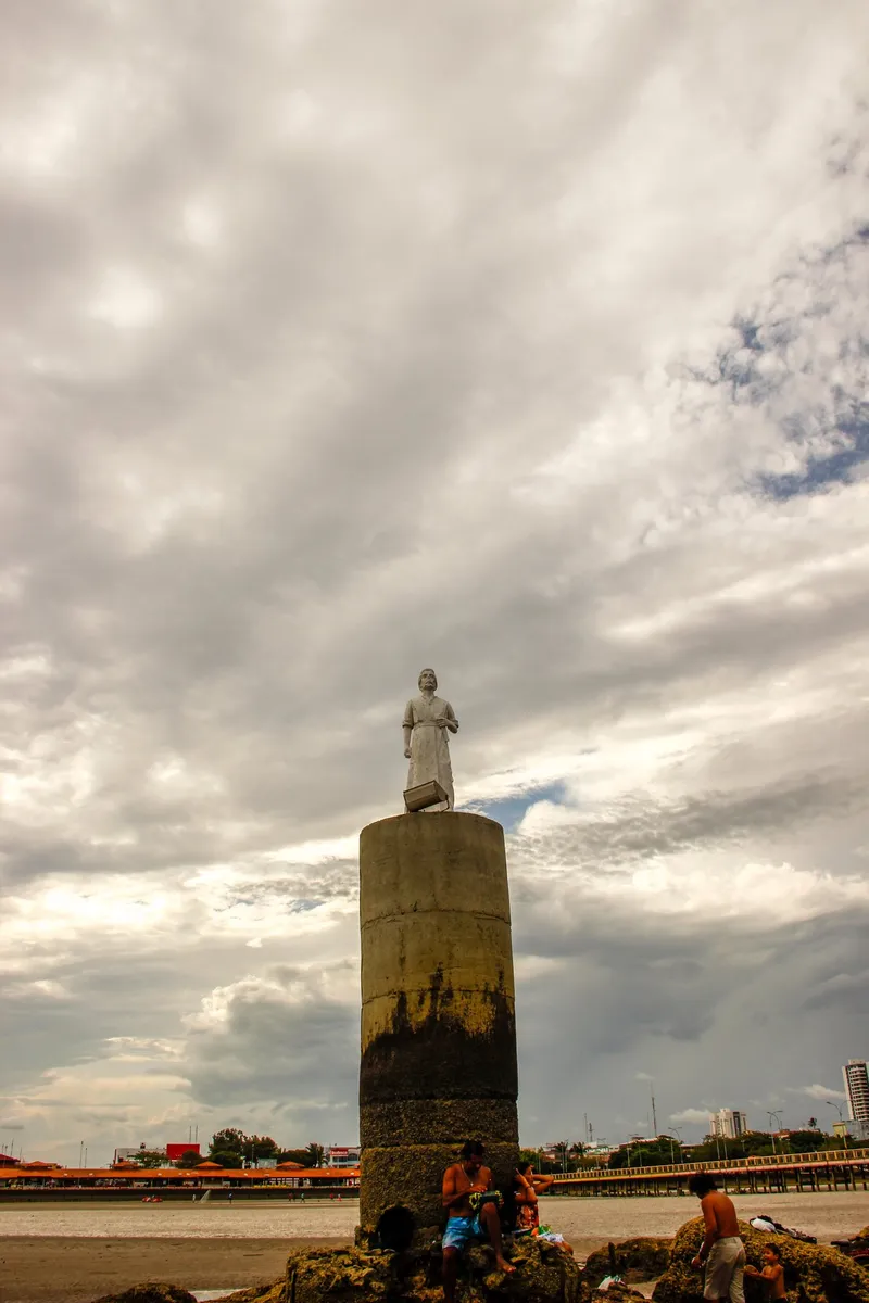 Imagem de São José sobre a coluna da Pedra do Guindaste vista de baixo, na maré baixa, com céu nublado e a orla de Macapá ao fundo.
