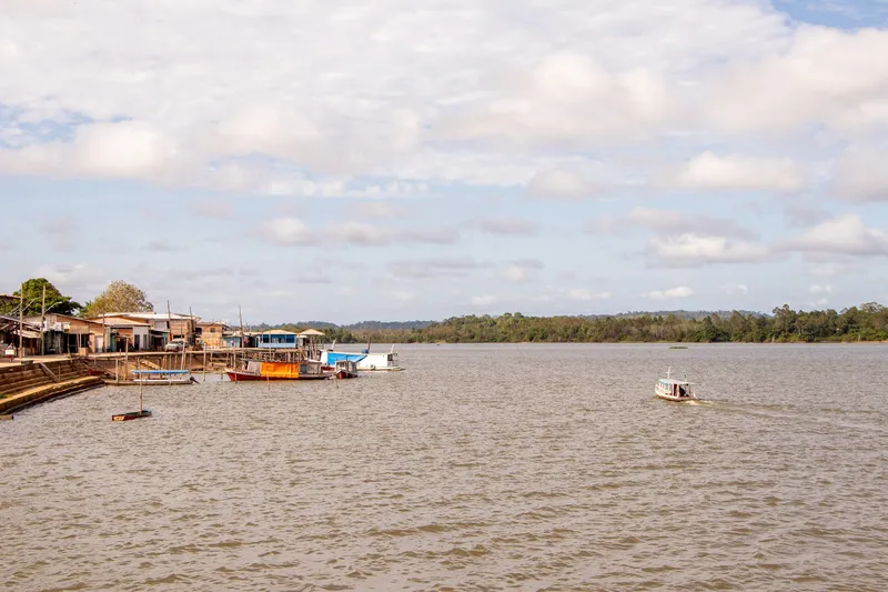 Wide river in front of the Vitória do Jari waterfront, with boats moored by the houses on the left and one boat underway, under a cloudy sky.