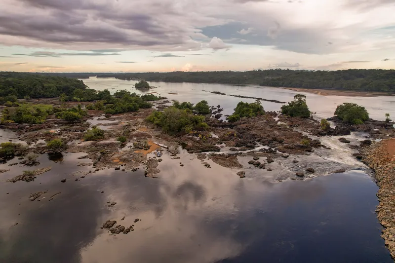 Vista aérea do rio Oiapoque na Corredeira Grand Roche ao entardecer, com rochas, ilhotas de mata e céu nublado refletido na água