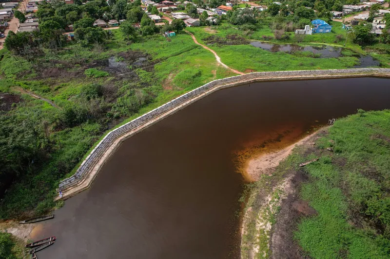 Aerial view of the Tartarugalzinho waterfront: a curved retaining wall along the dark-water river, with canoes on the bank and houses behind.