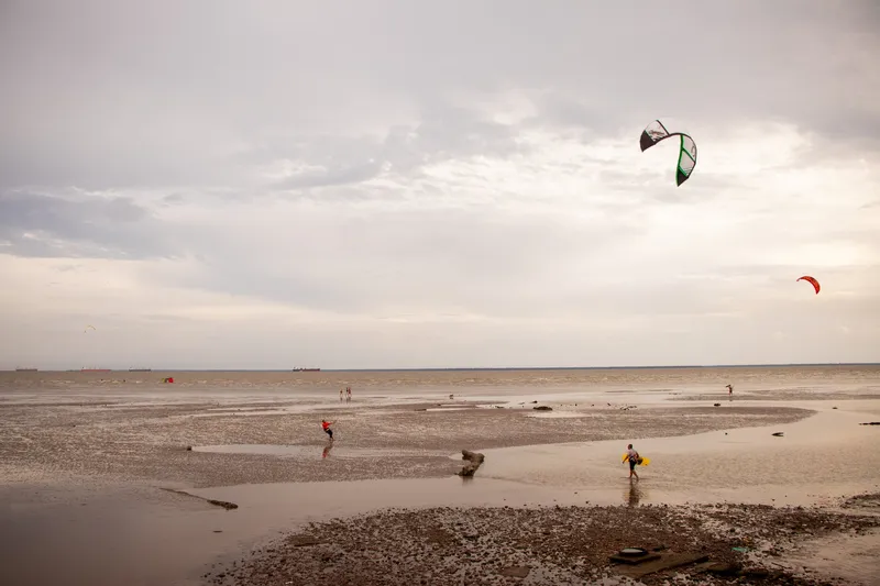 Praticantes de kitesurf na faixa de areia e lama da Orla do Bairro Cidade Nova na maré baixa, com pipas no céu nublado e navios no horizonte.