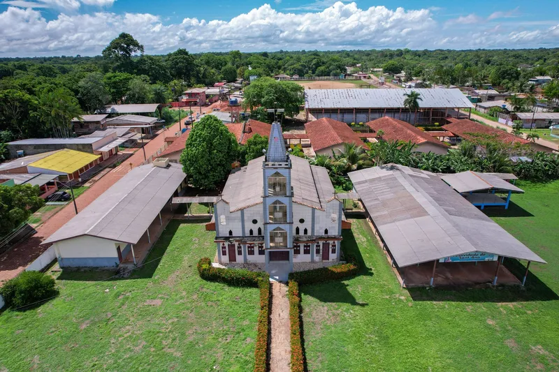 Aerial view of the Ilha de Santana church between covered sheds and lawn, with houses and forest around.