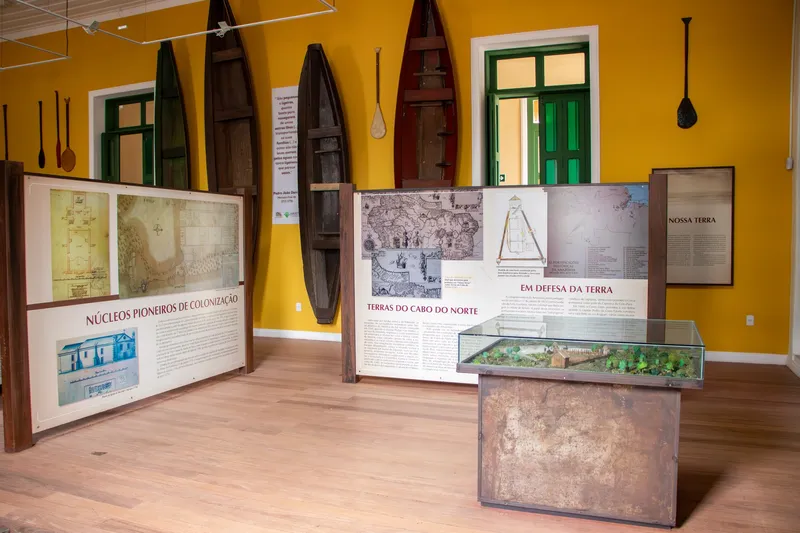Sala de paredes amarillas del Museu Joaquim Caetano da Silva con canoas y remos colgados, paneles con mapas antiguos y una maqueta en vitrina.